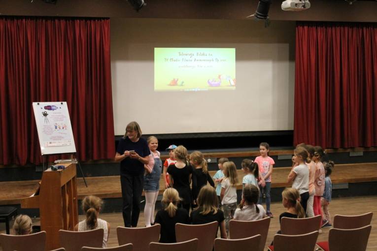 Auf dem Bild sieht man mehrere Kinder im Kreis stehen. Eine Frau schaltet Musik auf ihrem Telefon an. Im Hintergrund ist ein Plakat mit polnischer Schrift zu sehen.